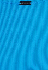 Tecido canelado em azul brilhante, com uma superfície texturizada. Uma pequena etiqueta preta com "WE ASH WE WEAR" está costurada na parte superior.