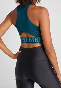 Turkusowy stanik sportowy z designem na plecy w kształcie racerback, z wycięciem. Dolna taśma jest elastyczna, z logo "PRO NIKE". Zestawiony z czarnymi legginsami.