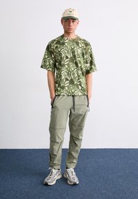 Green patterned short-sleeve shirt, olive cargo pants, and light gray sneakers. Beige cap with palm tree logo, black accents on pants.