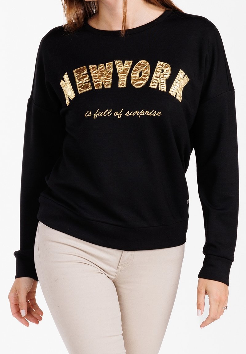 Femme portant un sweat-shirt noir à manches longues avec le texte doré "NEW YORK" et "is full of surprise" associé à un pantalon beige clair.