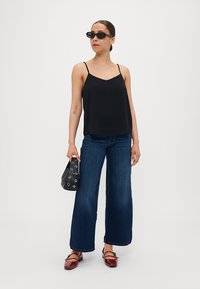 Svart satin camisole med tunna axelband, blå vida denimjeans och röda lackskor. Modellen håller en svart väska med silveröglor.