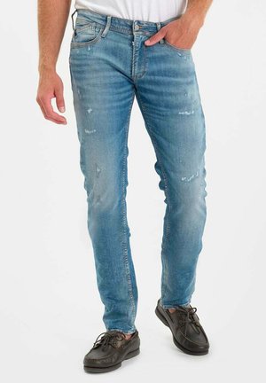 Man draagt lichtblauwe versleten smalvallende jeans en donkerbruine casual veterlaarzen, staande met één hand in de zak.