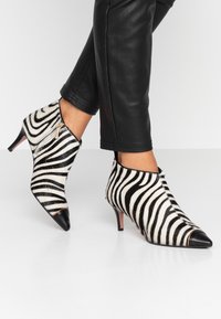 Zebra-print enkellaarzen van haar, met een zwarte puntige neus en een zijrits. De hak is laag en slank. Zwarte leren broek wordt gedragen.