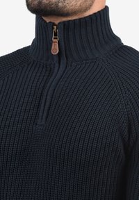 Dunkelblauer Strickpullover mit hohem Kragen, ausgestattet mit einem halben Reißverschluss und einem Lederanhänger. Gerippte Textur und strukturiertes Design.