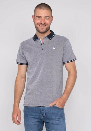 Homme souriant aux cheveux courts portant un polo gris à bordure noire et un jean bleu, debout avec une main dans la poche.