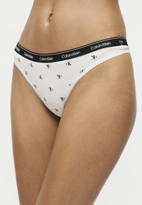 Γυναίκα που φοράει λευκά εσώρουχα Calvin Klein με μαύρη μέση και μικρά μαύρα λογότυπα διάσπαρτα στο ύφασμα.