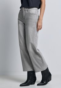 Persona che indossa jeans larghi grigio chiaro abbinati a stivaletti in pelle nera con tacco basso, in piedi davanti a uno sfondo neutro.