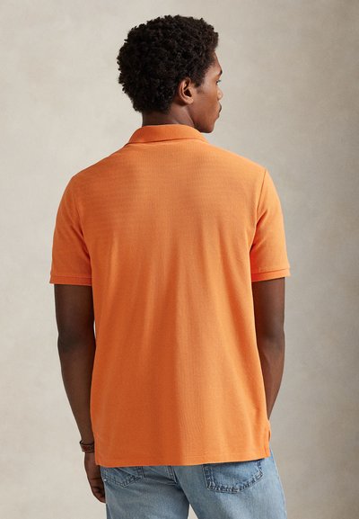 Πόλο με κοντά μανίκια σε έντονο πορτοκαλί χρώμα, κατασκευασμένο από απαλό ύφασμα με υφή. Διαθέτει κλασικό γιακά και πλαϊνές σχισμές στο τελείωμα.