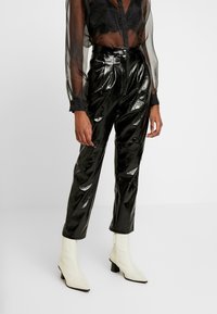 Černé lesklé vinylové kalhoty s vysokým pasem, zdobené záhyby a zúženými nohavicemi. Kombinovány s bílými špičatými kotníkovými botami.