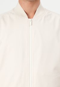 Light beige bomber jacket made of smooth fabric, featuring a stand-up collar, front zipper closure, and a subtle textured pattern.