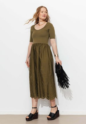 Femme portant une robe vert olive avec des détails en œillets, des sandales compensées noires, des bijoux en or et tenant un sac à main noir à franges.