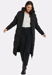 Sort dunjakke med hætte, der har et langt, quiltet design, lynlås fortil og trykknapper. Bæret over en lys top og mørke bukser med støvler.