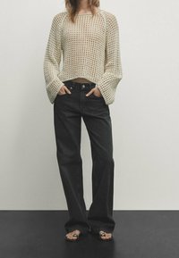 Helle beige, offen gestrickte Pullover mit langen, weiten Ärmeln, kombiniert mit hoch taillierten, dunkelgrauen, weiten Jeans und flachen Sandalen.