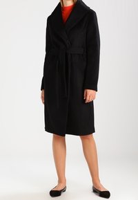 Schwarzer Mantel mit Gürtel und großem Kragen, aus weichem Material gefertigt. Verfügt über lange Ärmel und einen mittellangen Schnitt. Wird über einem roten Oberteil getragen.