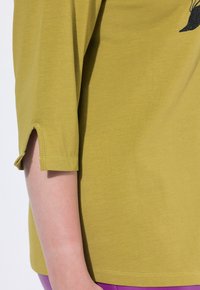 Camisa verde oliva con mangas tres cuartos, que presenta una abertura en el puño. Tela de algodón suave con una textura lisa y un diseño casual.