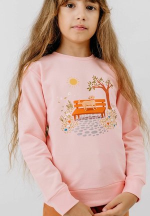 Young girl with long hair wearing a pink sweatshirt featuring a park scene with a dog on an orange bench, tree, sun, and flowers.