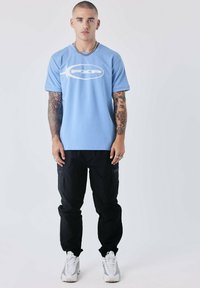 Camiseta de algodón azul claro con un logo blanco, combinada con pantalones cargo negros con cremalleras y zapatillas blancas con detalles grises.