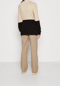 Person som bär en långärmad tröja i beige och svart, beige vida byxor och vita sneakers med gula sulor, står vänd bort.