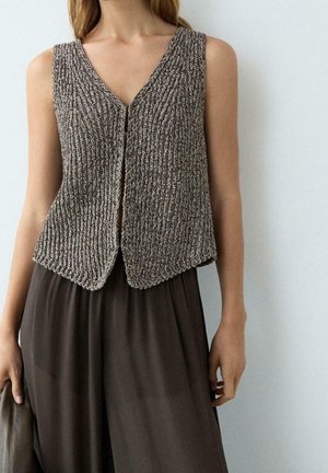 Femme portant un gilet tricoté marron et blanc sans manches, par-dessus un pantalon ample marron foncé, debout contre un mur clair uni.