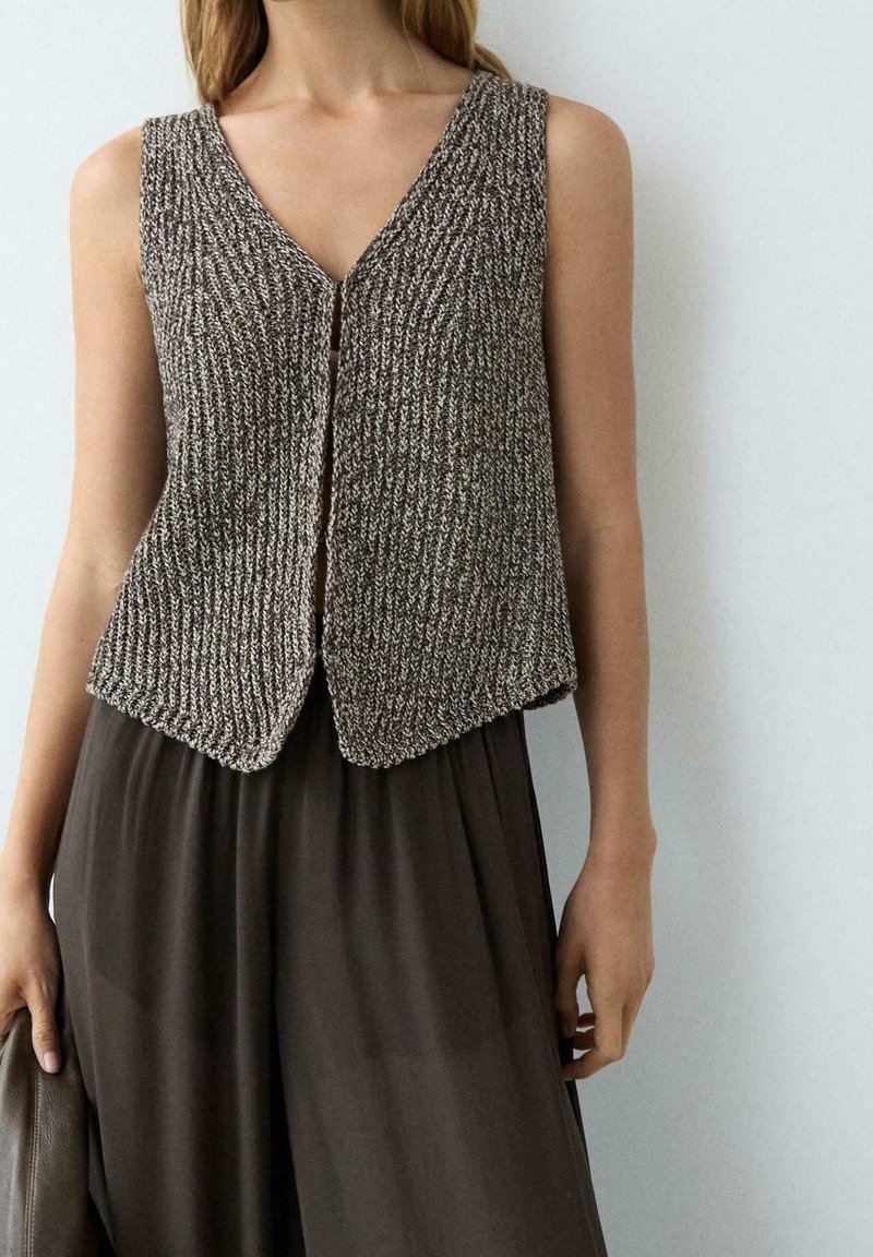 Femme portant un gilet tricoté marron et blanc sans manches, par-dessus un pantalon ample marron foncé, debout contre un mur clair uni.