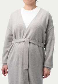 Ljusgrå, lång cardigan gjord av mjukt material, med omslagdesign, bälte och vida ärmar. Bärs över en vit skjorta.