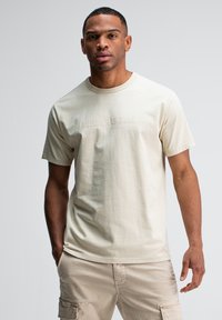 Man in beige cargoshorts en een off-white t-shirt met korte mouwen met subtiele tekst, staande tegen een effen lichtgrijze achtergrond.