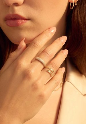 Goldringe mit glatten und strukturierten Oberflächen, die an einer Hand getragen werden. Die Nägel sind hellrosa; die Haut ist hell. Der Schmuck ist minimal und betont die Einfachheit.