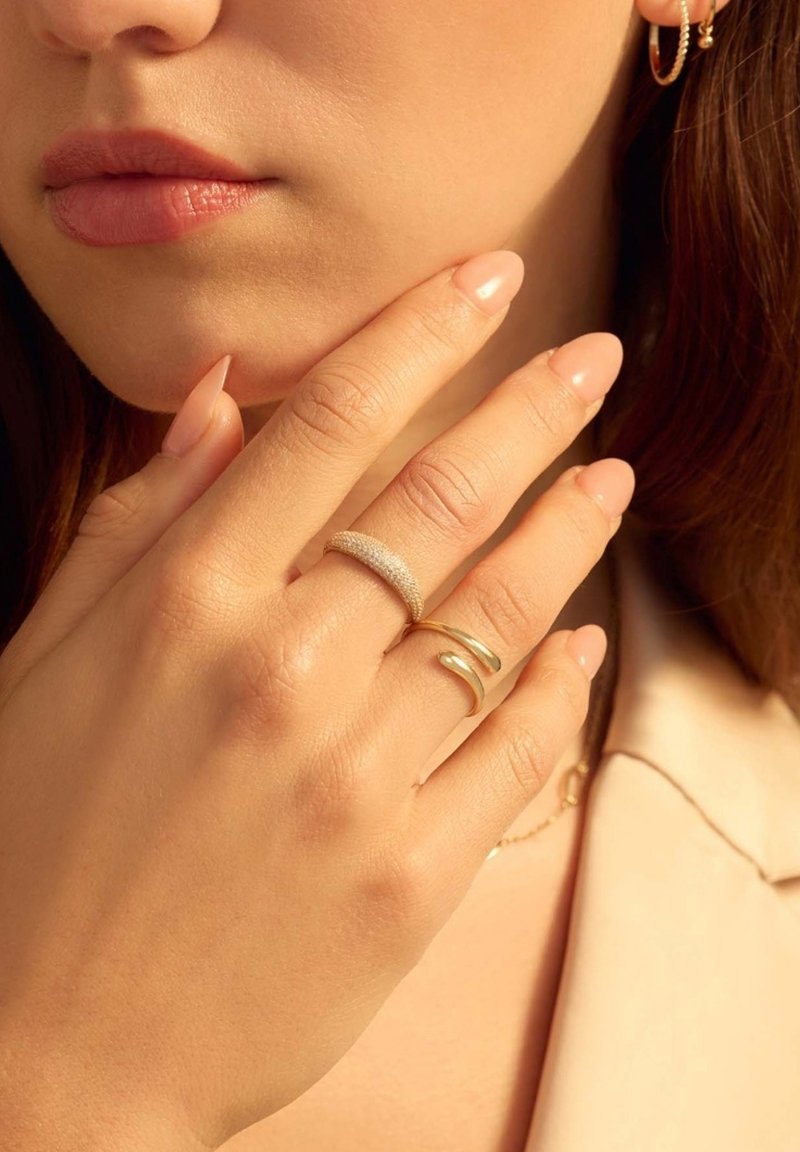 Goldringe mit glatten und strukturierten Oberflächen, die an einer Hand getragen werden. Die Nägel sind hellrosa; die Haut ist hell. Der Schmuck ist minimal und betont die Einfachheit.