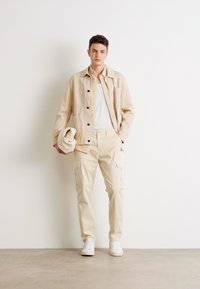 Chaqueta de algodón beige con botones y bolsillos, combinada con pantalones cargo beige que tienen bolsillos laterales, usados con una camiseta blanca y zapatillas.