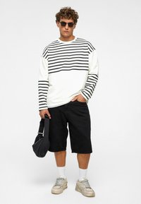 Wit en zwarte gestreepte lange mouwen shirt, zwarte shorts, grijze sneakers en een zwarte schoudertas. Model draagt een zonnebril en heeft krullend haar.