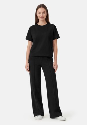 Nuori nainen seisoo, pukeutunut yksinkertaiseen mustaan lyhythihainen paita, mustiin leveälahkeisiin housuihin ja valkoisiin lenkkareihin valkoista taustaa vasten.