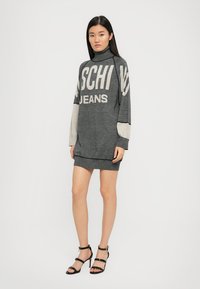 Pelēka adīta kleita ar augstu kakla izgriezumu, pārmērīgu piegriezumu, kontrastējošām piedurknēm un izteiksmīgu tekstu "MOSCHINO JEANS" gaiši pelēkā krāsā. Akcentēta ar apakšmalu.