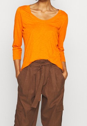 Kobieta w jaskrawo pomarańczowej bluzce z długim rękawem, wpuszczonej w spodnie z wysokim stanem w kolorze brązowym, z materiałowym paskiem, z rękami w kieszeniach.