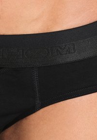 Mutande in cotone nere con una cintura elastica texturizzata che presenta un logo. Il tessuto è liscio, con un design aderente e dettagli di cucitura visibili.