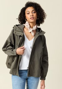Giacca con cappuccio verde oliva, con zip frontale, polsini elasticizzati e cordini. Abbinata a una maglietta bianca a scollo a V e jeans di denim.