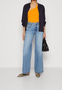 Orange one-shoulder top, navy bomber jacket, high-waisted light blue wide-leg jeans, olive green flats, and a black handbag.