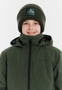 Grüne wasserdichte Jacke mit hohem Kragen und schwarzem Reißverschluss, kombiniert mit einer grünen gerippten Strickmütze mit einem Logo-Patch.