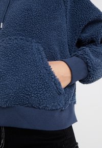 Sudadera de forro polar en azul oscuro, con un cuerpo texturizado, puños acanalados y un bolsillo frontal. La tela suave contrasta con la superficie esponjosa.