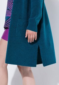 Teal stickad cardigan med texturerad yta, ribbade manschetter och en sidoklyvning. Har en enkel ficka fram.