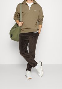 Sweatshirt zippé vert olive, T-shirt blanc en dessous, pantalon en velours côtelé marron et baskets blanches. Porte un sac vert.