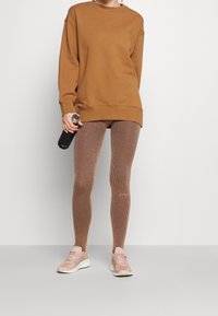 Sudadera marrón con textura acanalada, combinada con leggins marrones a rayas. Se sostiene una botella de agua negra y lleva zapatillas deportivas beige.