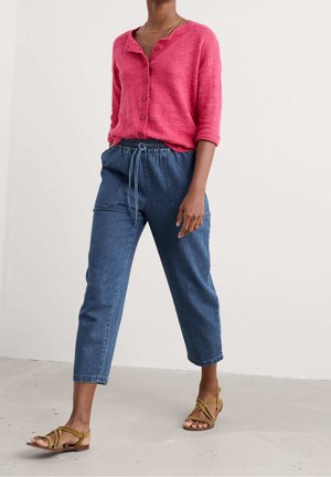 Kobieta w jasnoróżowym kardiganie zapinanym na guziki, niebieskich dżinsowych spodniach o podwyższonym stanie z nogawkami do połowy łydki ze ściągaczem oraz w brązowych, płaskich sandałach na paski, idąca spacerem.