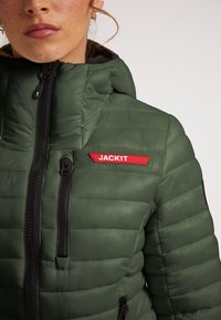 Grøn puffy jakke med høj krave, sorte lynlåse og et rødt logo patch mærket "JACK1T" på brystet. Tekstureret, quiltet design.
