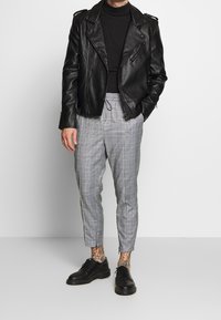 Man wearing black leather jacket, black turtleneck, gray plaid cropped pants, black shoes, and visible ankle tattoos, standing against white wall.