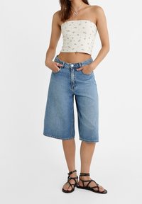 Mujer vistiendo un top corto sin mangas blanco con flores, pantalones cortos de denim holgados hasta la rodilla, sandalias negras con tiras y un collar en forma de corazón.