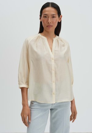 Femme portant un chemisier ample couleur crème avec des manches trois-quarts et un jean bleu clair, debout devant un fond uni.