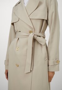 Beige double-breasted trench coat with gold buttons, featuring a tied belt, notched lapels, and adjustable cuffs. Smooth texture.