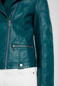 Turkusowa skórzana kurtka motocyklowa z zamkiem błyskawicznym z przodu, boczną kieszenią i teksturowanymi przeszyciami na rękawach. Akcenty ze srebrnego metalu.