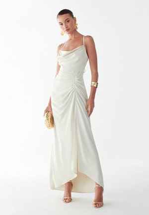 Kobieta w kremowej, marszczonej sukni wieczorowej na cienkich ramiączkach, trzymająca złotą kopertówkę, nosząca złote kolczyki, bransoletkę oraz sandały na cienkich paskach.