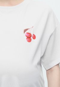 Λευκό μπλουζάκι με ένα μικρό γραφικό τριών κόκκινων κερασιών και ένα καφέ φύλλο στην πάνω αριστερή πλευρά του στήθους.